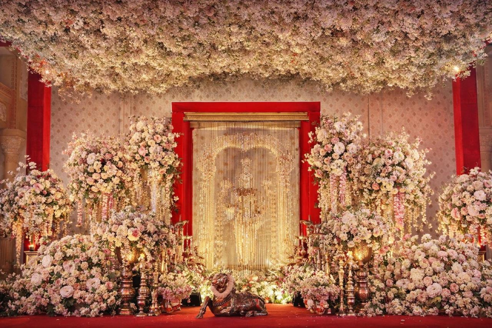 Blush floral ceiling and golden bead curtain backdrop with brass nandi at a temple-inspired wedding stage — Panigrahana decor
