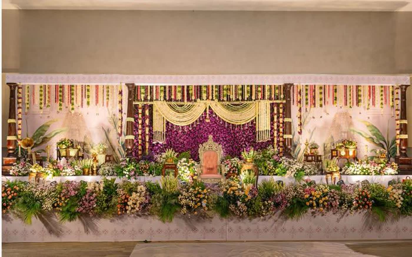 Long reception stage with a purple floral centre panel, gold lanterns, palm and mixed seasonal florals along the full front edge