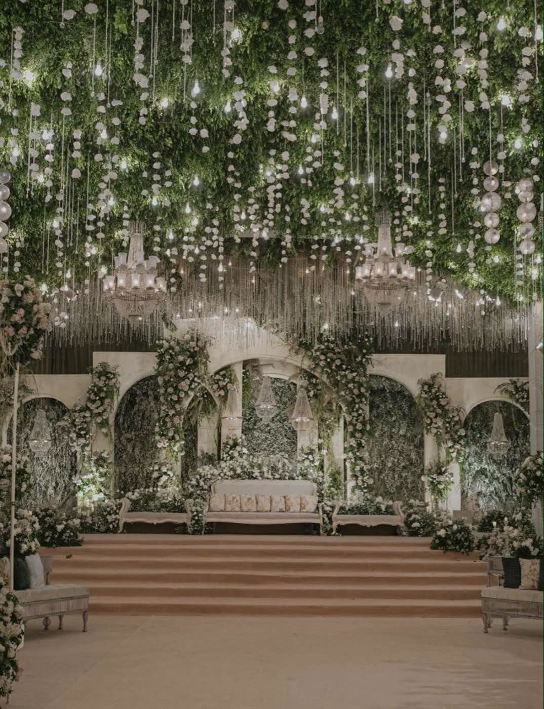 Arched reception backdrop in white roses and greenery under a suspended canopy of hanging bulbs and crystal chandeliers