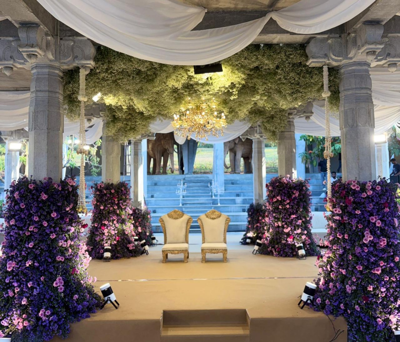 Stone-pillared pavilion dressed in purple and pink floral columns under a baby's breath ceiling and chandelier — Panigrahana decor