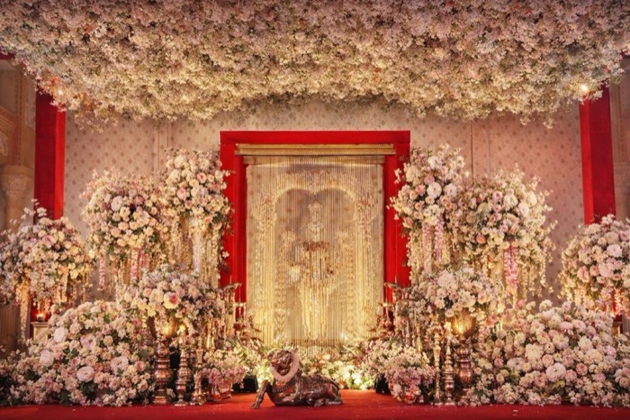 Blush floral ceiling and golden bead curtain backdrop with brass nandi at a temple-inspired wedding stage — Panigrahana decor