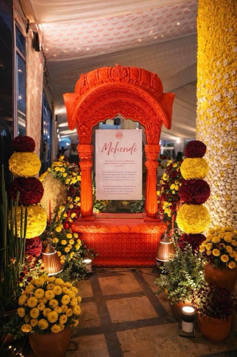 Carved orange jharokha frame with marigold topiaries marking the mehendi entrance — Panigrahana decor