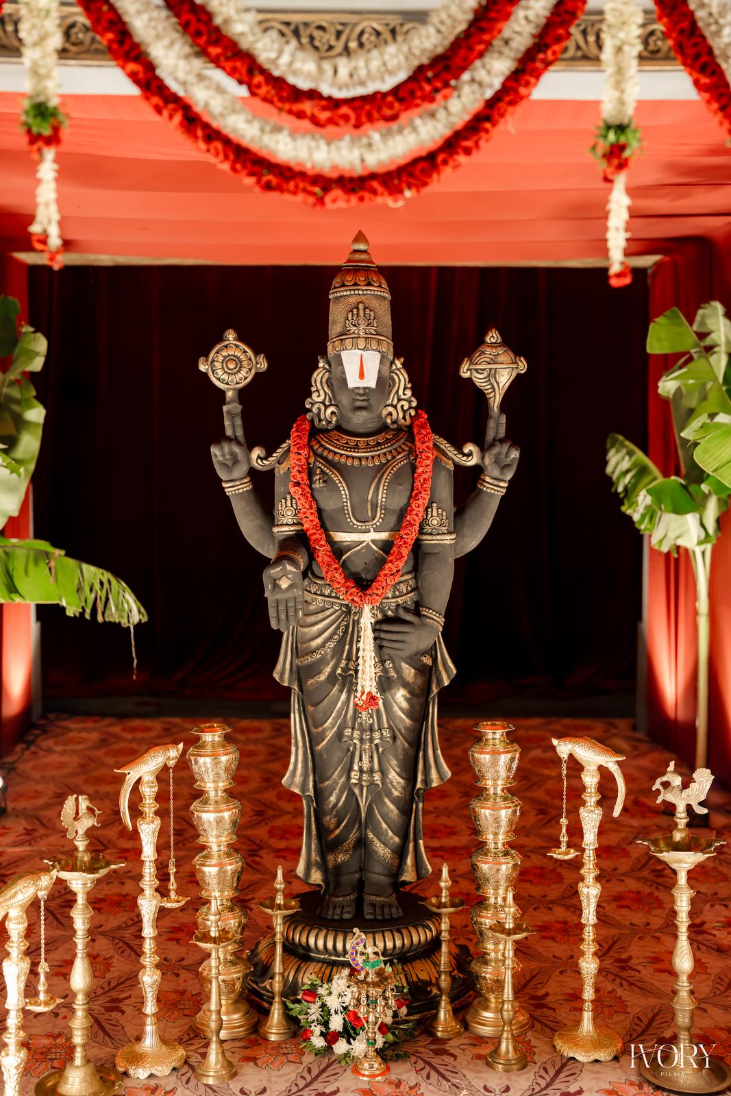 Garlanded deity sculpture flanked by rows of tall brass lamps