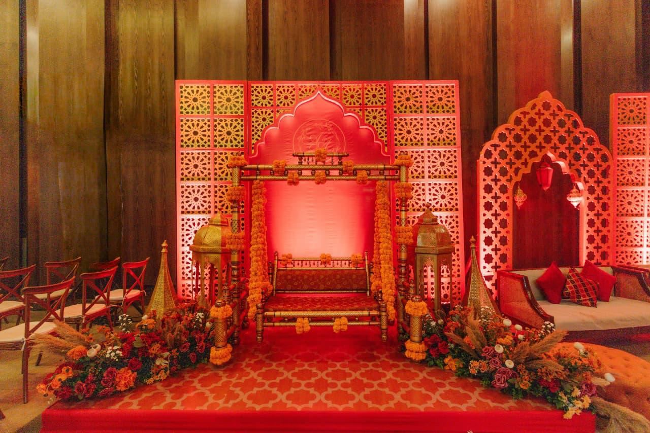 Red jharokha-style stage with carved arches, lanterns and floral beds