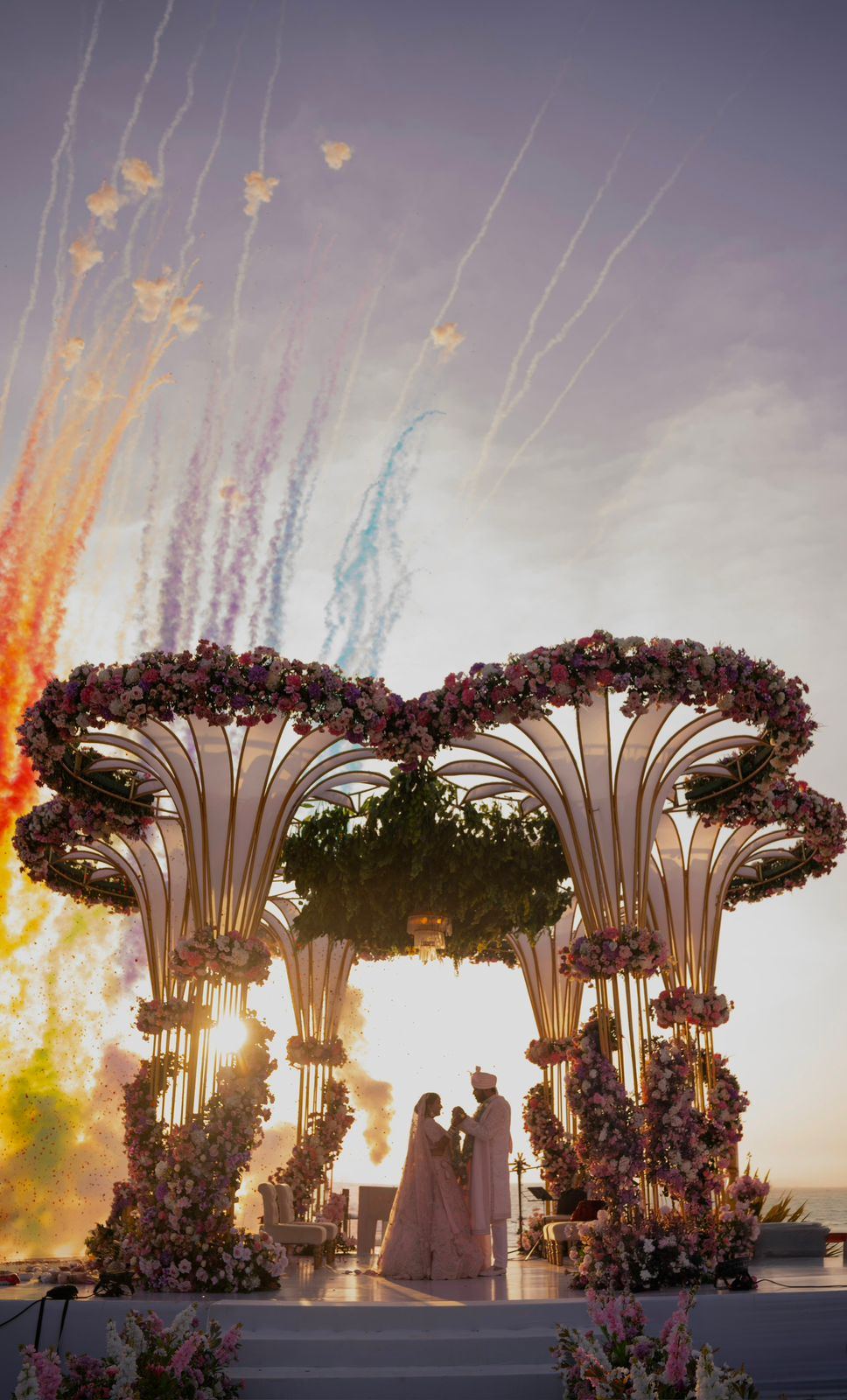 Couple beneath a sculptural floral mandap silhouetted against the sunset sky