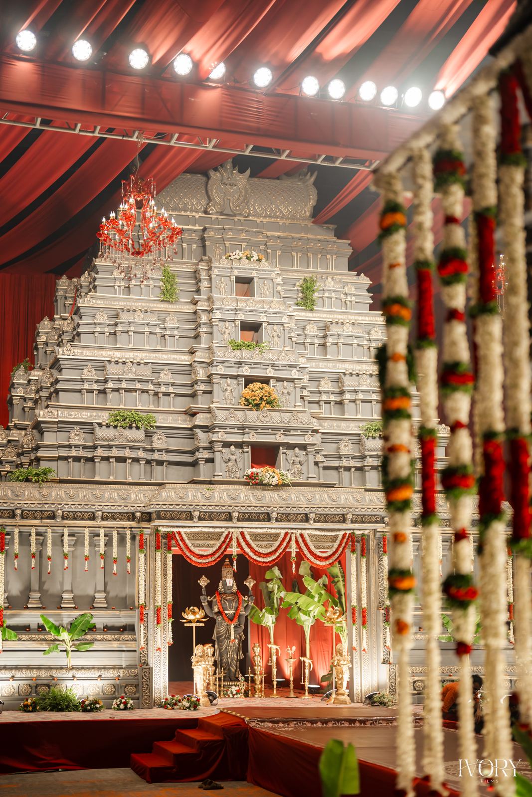 Carved gopuram-style mandap replica framed by red drapes and floral pillars