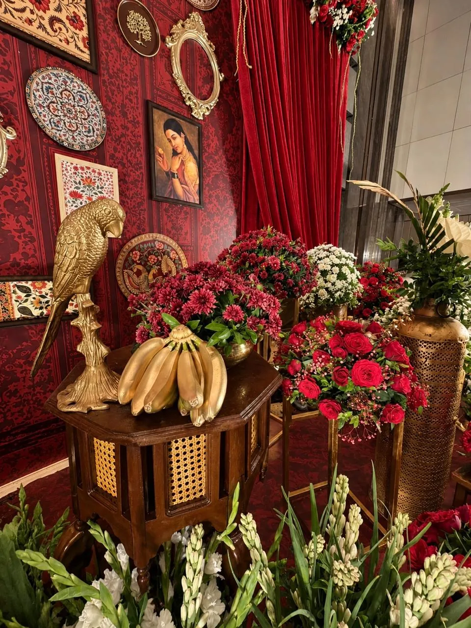 A brass parrot and a bunch of gold-painted bananas on a carved cane side table, with burgundy chrysanthemums and red roses against the red damask stage wall