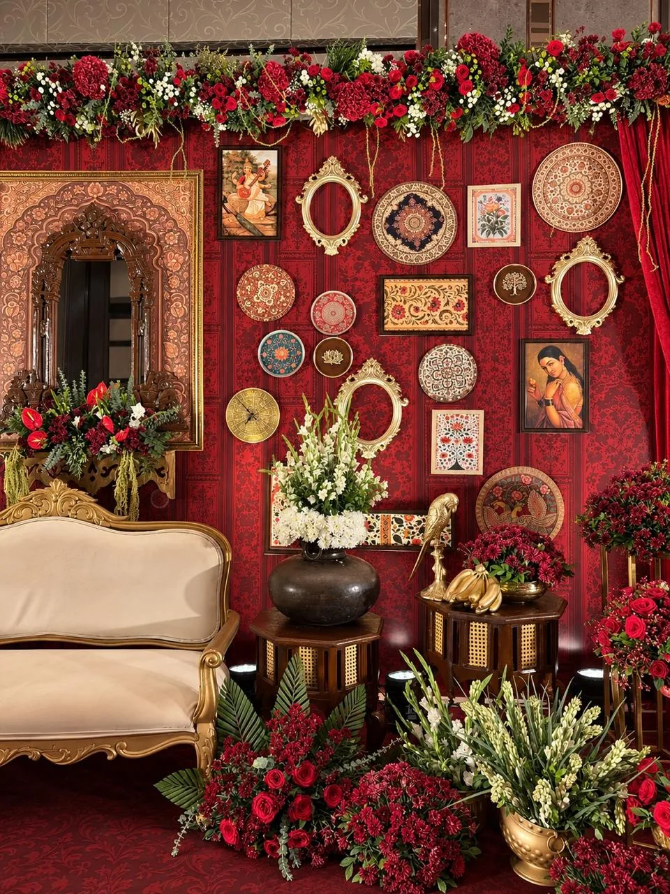 Detail of the half saree stage wall: a Raja Ravi Varma Saraswati print, Mughal-style floral frames, hand-painted plates, empty gilded frames, a carved mirror above a gilded settee, and tuberose in a black clay pot