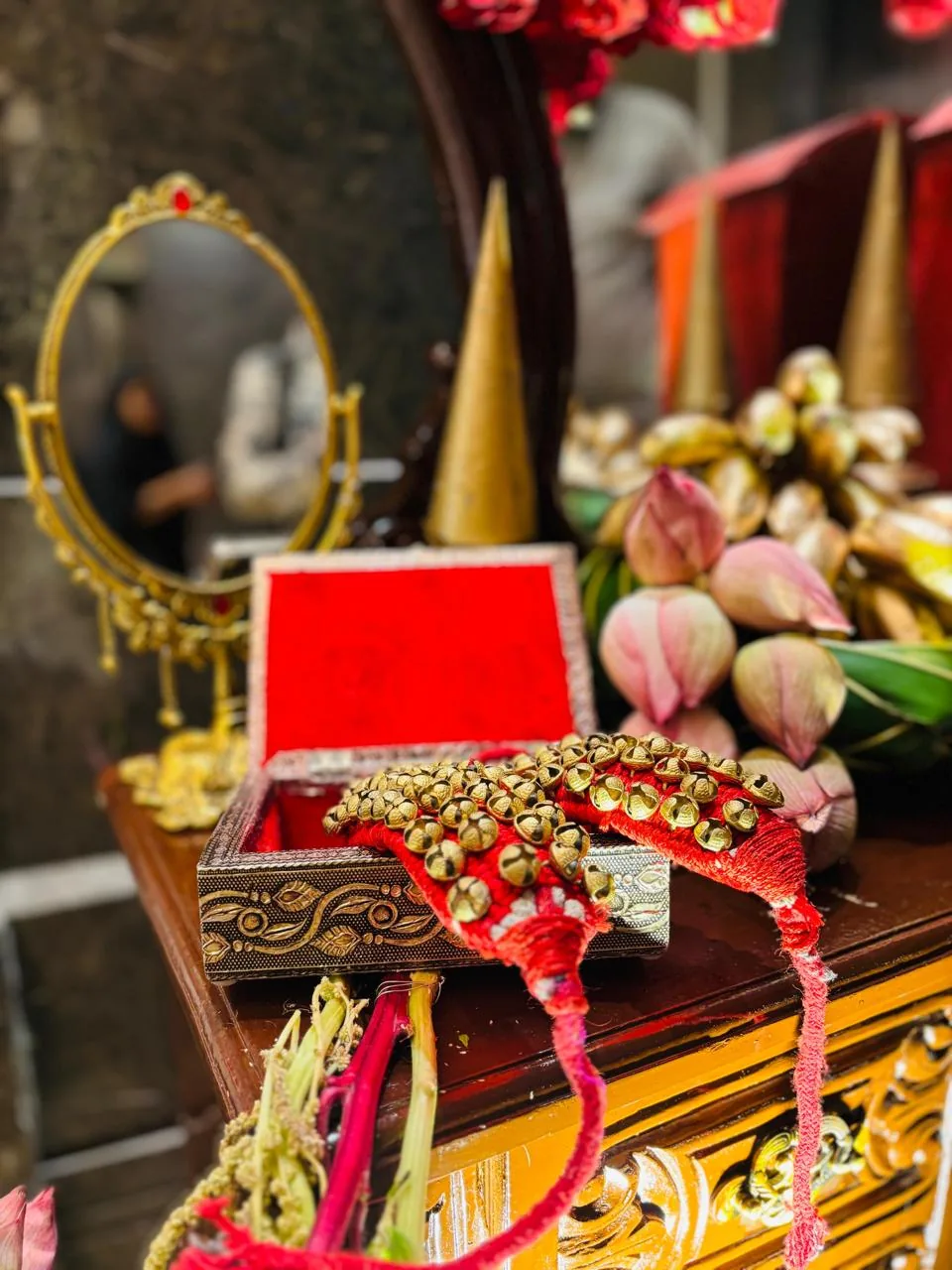 Ghungroo anklet bells on red cord in a metal-and-velvet box, with a gold vanity mirror, gold cones and pink lotus buds, styled for a half saree ceremony