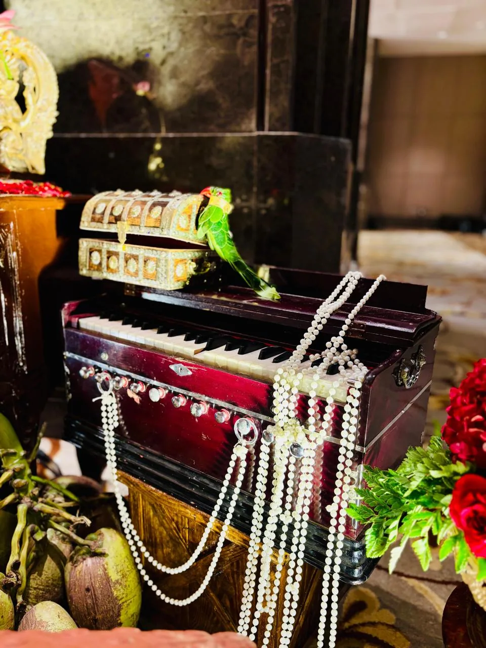 A harmonium draped in pearl strings beside a brass-inlaid trunk topped with a green parrot, tender coconuts and red roses, part of the half saree ceremony styling