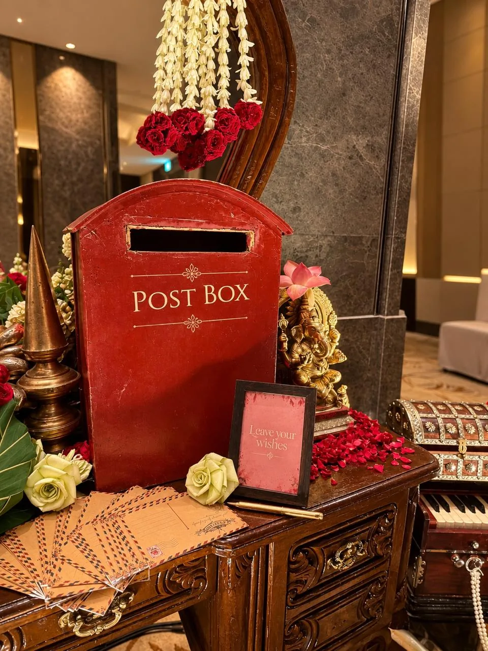 A red post box for guests' wishes with a 'Leave your wishes' card, airmail postcards, a brass Ganesha, a brass cone and jasmine with red roses on a carved desk