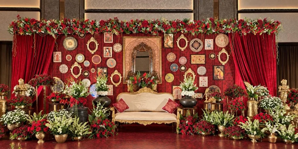 Half saree ceremony stage at Artezia Bengaluru (formerly Shangri-La): a red damask gallery wall of Raja Ravi Varma prints, painted plates and gilded frames around a carved mirror, a gilded settee, red velvet drapes, brass lamp-bearer statues and red rose and tuberose arrangements