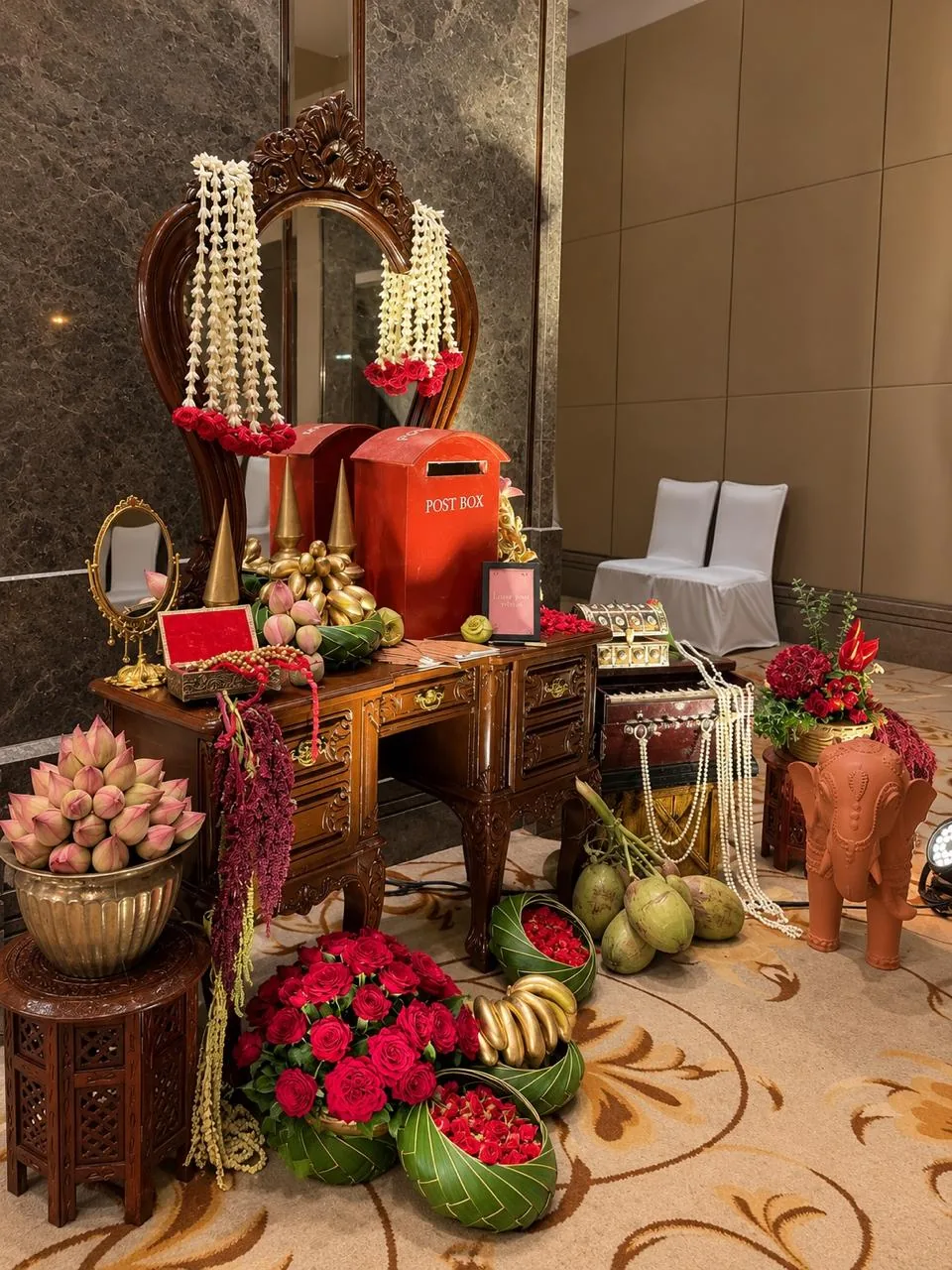 Vintage wishing desk for guests: a carved dressing table and mirror garlanded with jasmine and roses, red post boxes, pink lotus buds in a brass urli, a harmonium draped in pearls, tender coconuts, gold-painted bananas and a terracotta elephant