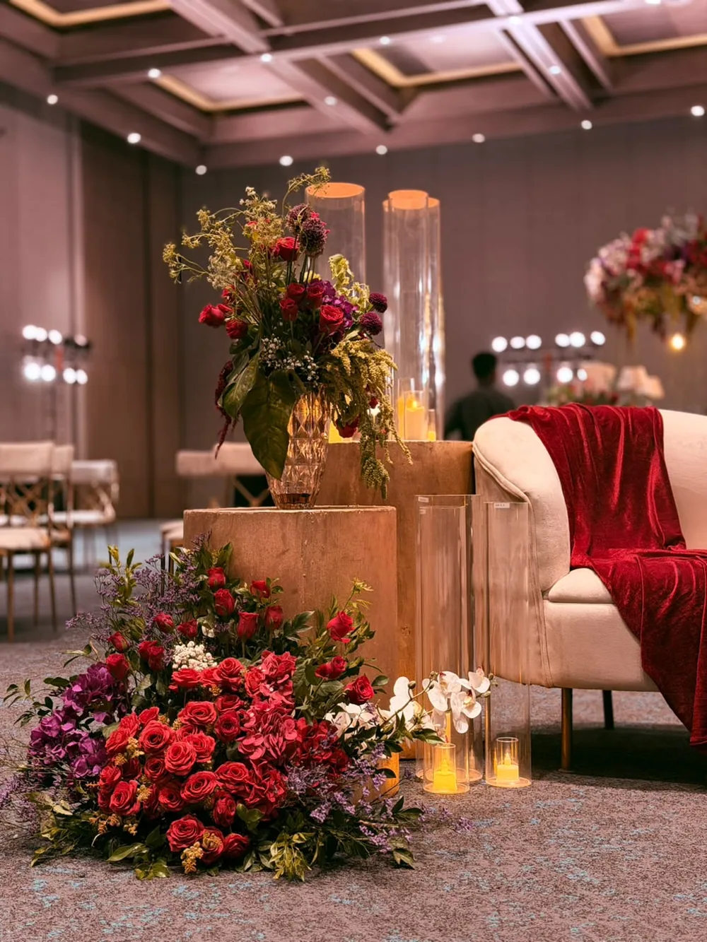Red roses, magenta bougainvillea and white orchids banked at a gold plinth beside a cream lounge and a red velvet throw, under the coffered ballroom ceiling at JW Marriott Bengaluru Prestige Golfshire. Panigrahana Weddings.
