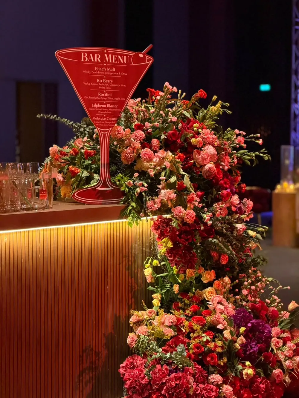 A cocktail-glass bar menu standing in a fall of coral, red and pink carnations and spray roses down the front of a lit bar at JW Marriott Bengaluru Prestige Golfshire. Panigrahana Weddings.