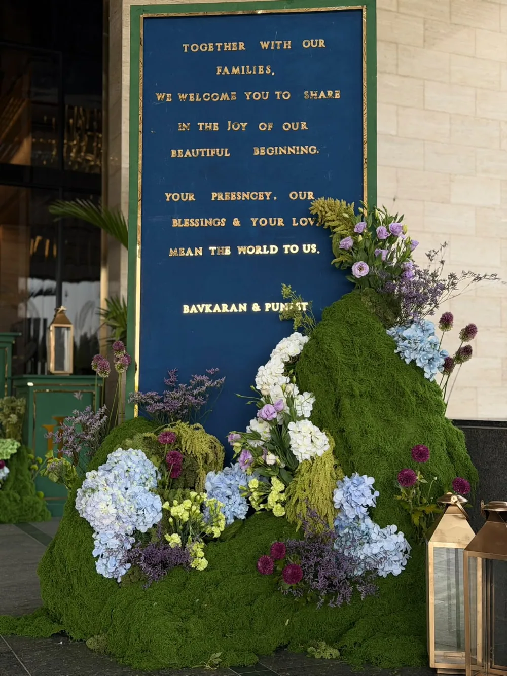A blue and gold welcome board framed by a moss bank of hydrangea, allium, amaranthus and dried limonium at JW Marriott Bengaluru Prestige Golfshire. Panigrahana Weddings.