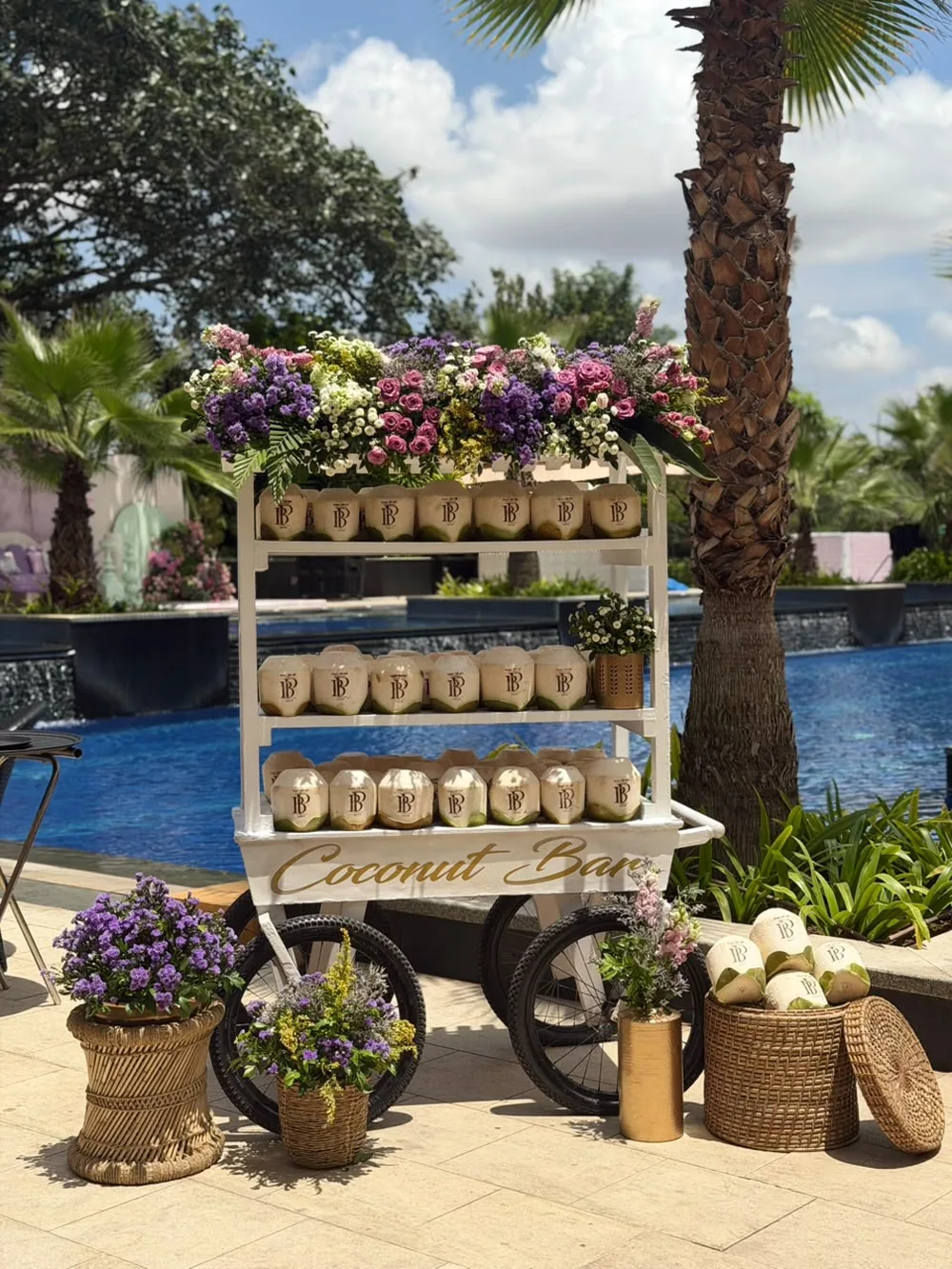 A white coconut bar cart topped with pink and lilac stock, roses and limonium, stacked with monogrammed coconuts beside the pool at JW Marriott Bengaluru Prestige Golfshire. Panigrahana Weddings.