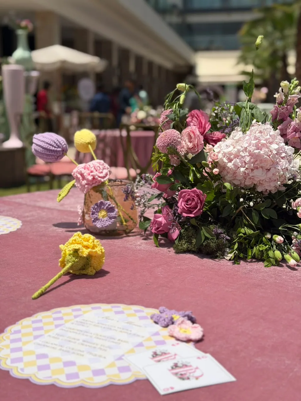 Hand-crocheted flowers, a lilac and yellow checkerboard menu card and playing cards on rose velvet, beside pink hydrangea and garden roses at JW Marriott Bengaluru Prestige Golfshire. Panigrahana Weddings.