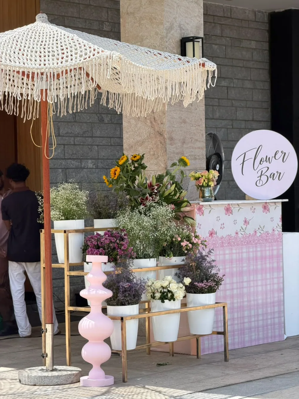 A flower bar of white buckets filled with gypsophila, limonium, lilies and sunflowers under a fringed macramé parasol at JW Marriott Bengaluru Prestige Golfshire. Panigrahana Weddings.
