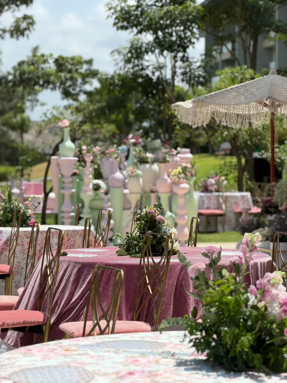 Round tables dressed in rose velvet and floral chintz with gold chairs and low garden arrangements, under an avenue of pastel urns at JW Marriott Bengaluru Prestige Golfshire. Panigrahana Weddings.