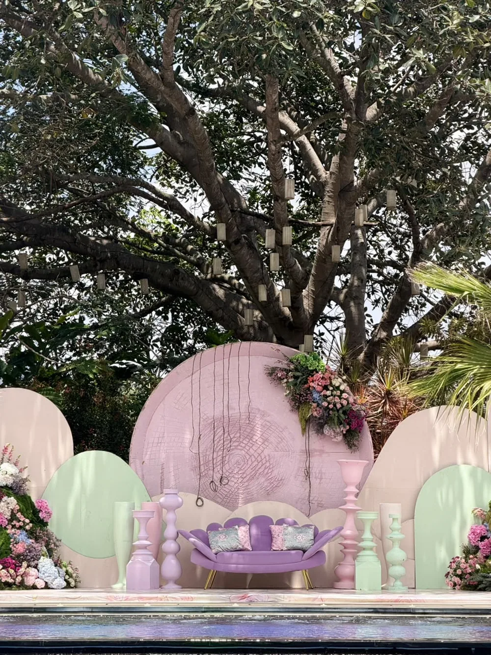 A lilac scalloped sofa on the pastel poolside stage beneath a rain tree hung with paper tassels at JW Marriott Bengaluru Prestige Golfshire. Panigrahana Weddings.