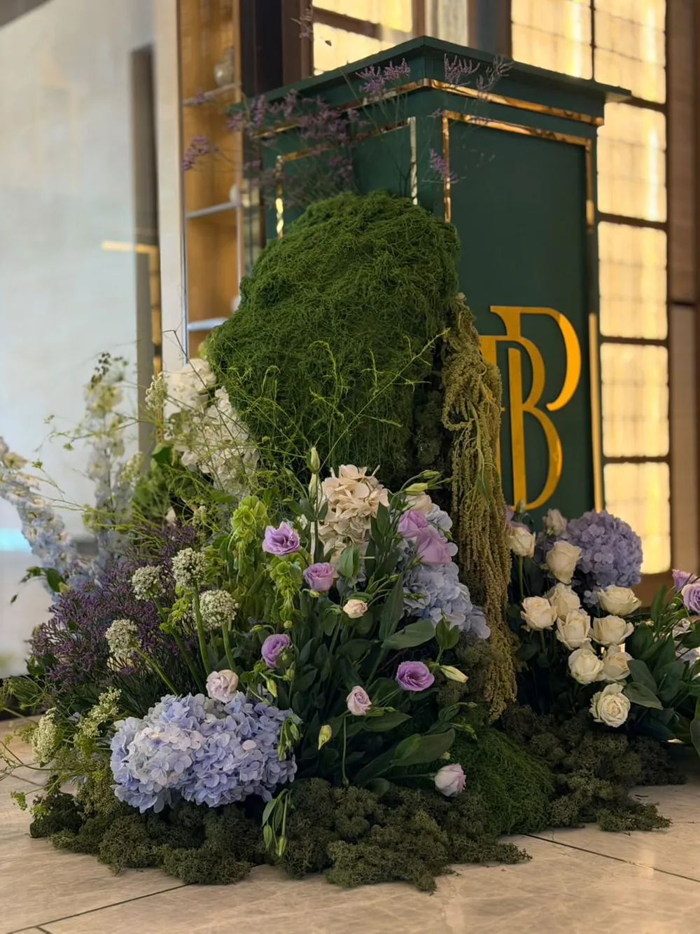 An indoor moss column beside a green and gold monogram plinth, planted with lilac lisianthus, blue hydrangea, ivory roses and trailing amaranthus at JW Marriott Bengaluru Prestige Golfshire. Panigrahana Weddings.