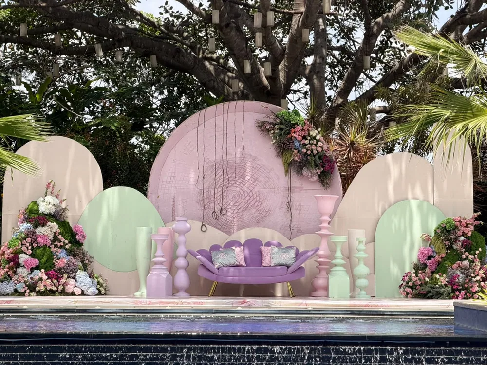 The full mehendi stage in blush and mint arches with turned pastel urns, floral mounds and a lilac sofa, mirrored in the pool at JW Marriott Bengaluru Prestige Golfshire. Panigrahana Weddings.