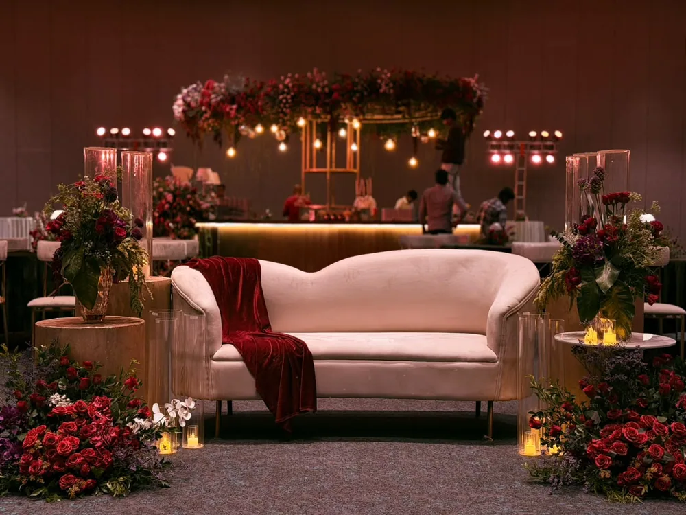 A blush lounge with a red velvet throw between red rose mounds, under a suspended circular floral chandelier hung with warm bulbs over the bar at JW Marriott Bengaluru Prestige Golfshire. Panigrahana Weddings.
