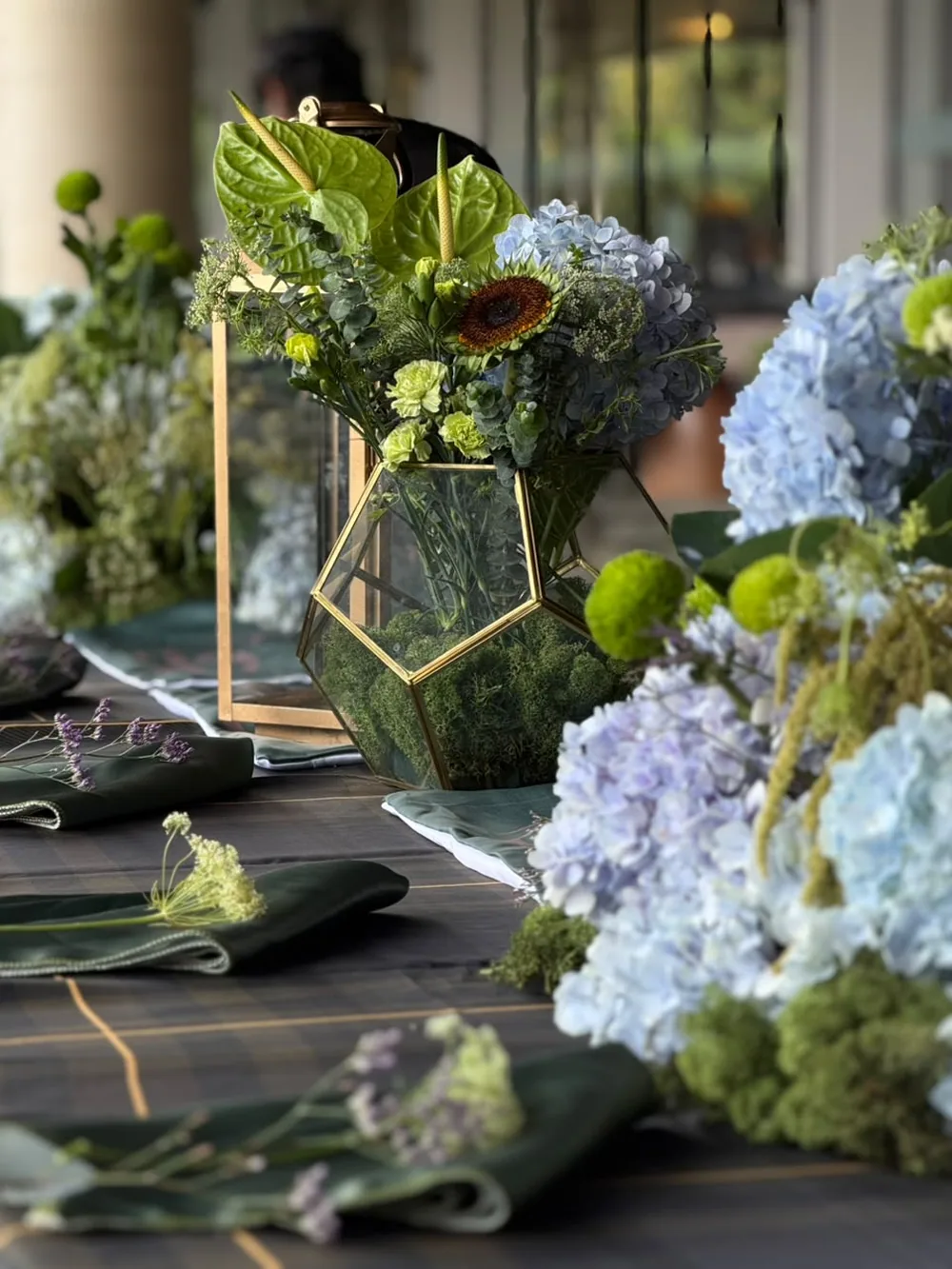 A gold geometric terrarium packed with moss, set among blue hydrangea, green anthurium and a single sunflower on a tartan table with deep-green napkins at JW Marriott Bengaluru Prestige Golfshire. Panigrahana Weddings.