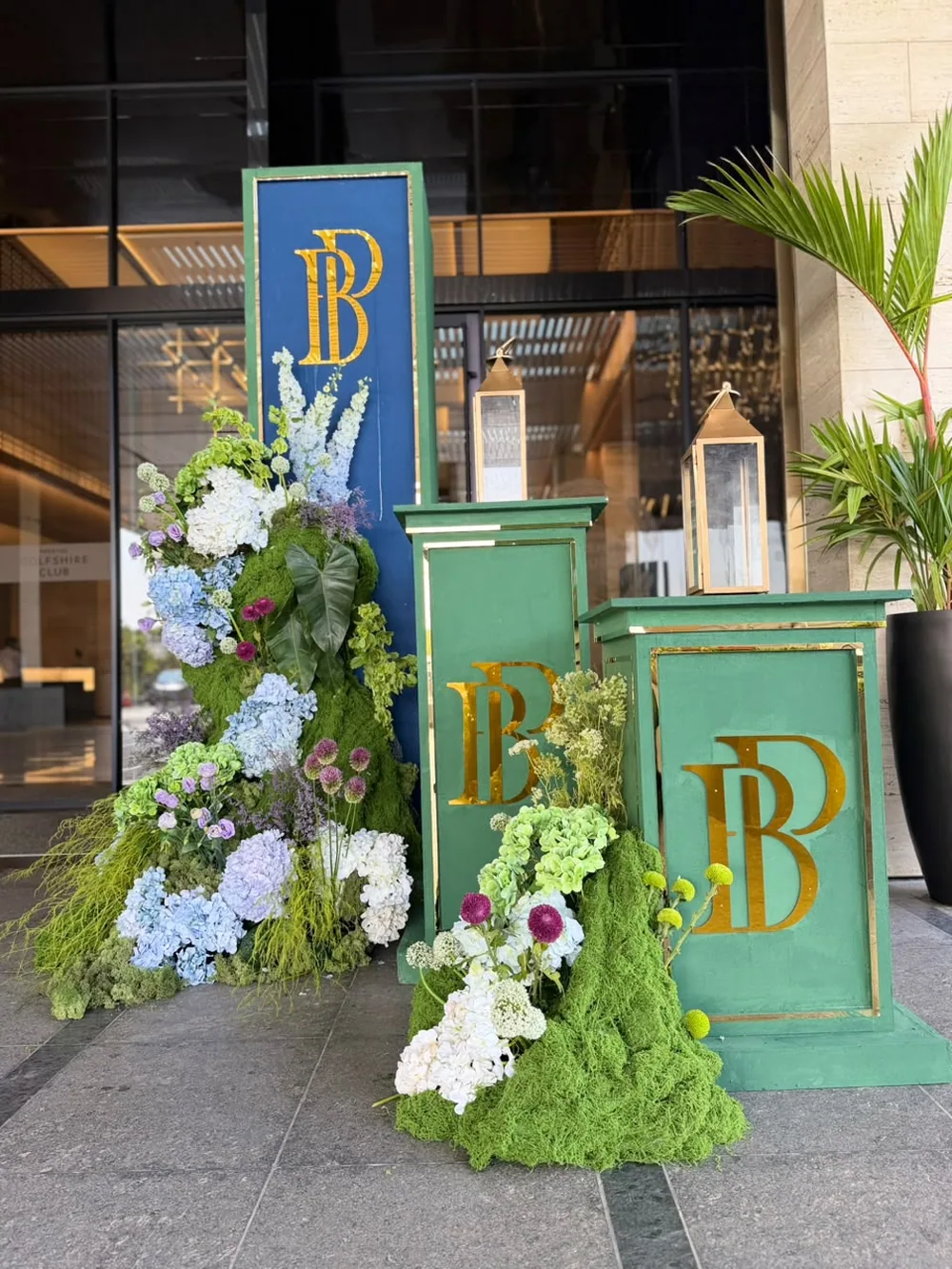 Moss-clad plinths carrying a gold monogram, banked with blue and white hydrangea, lisianthus and allium at the entrance to JW Marriott Bengaluru Prestige Golfshire. Panigrahana Weddings.