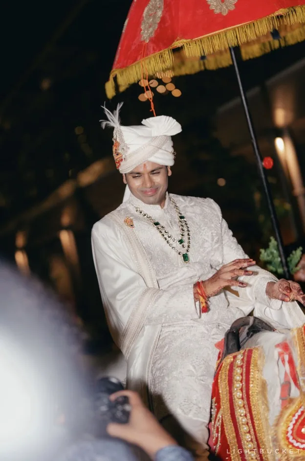 Groom in an ivory sherwani arriving for the baraat under a red and gold embroidered umbrella at Nature Knots, Bangalore