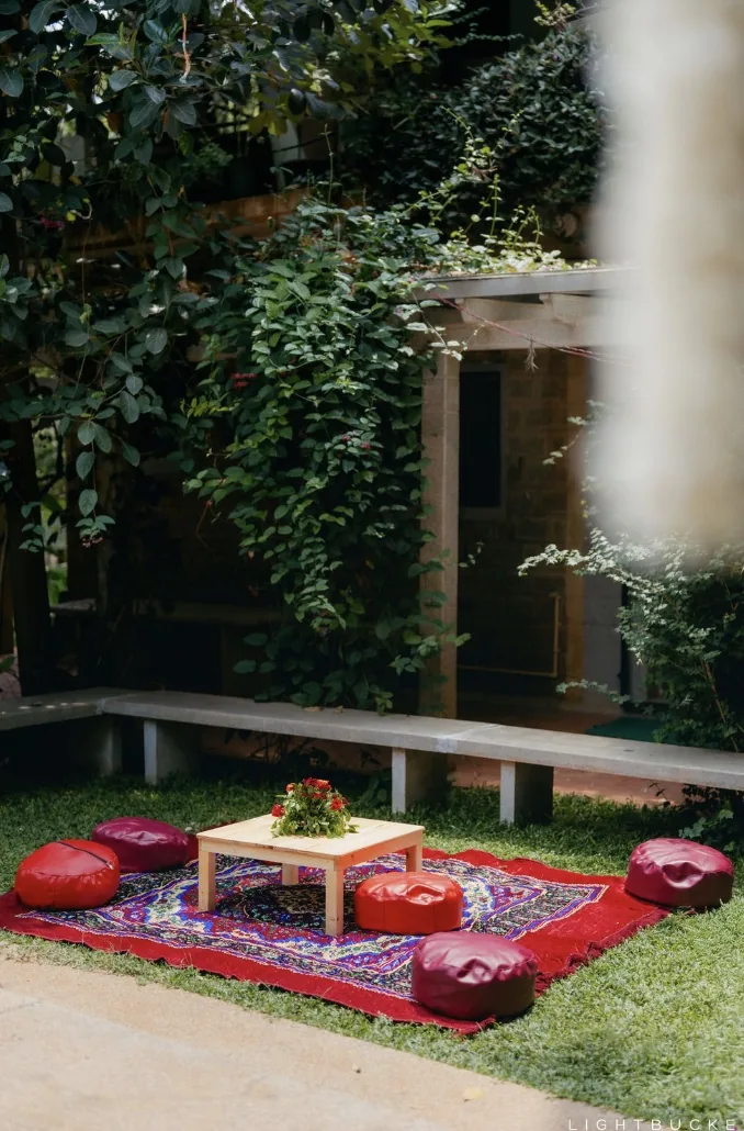 Low floor seating on the lawn at Nature Knots, Bangalore: red leather poufs and a wooden table on a red patterned rug under the trees