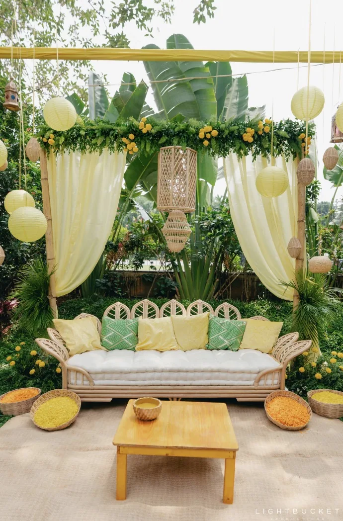 Haldi lounge with a cane sofa and yellow and green cushions under a frame of sheer lemon drapes, greenery and lanterns at Nature Knots, Bangalore