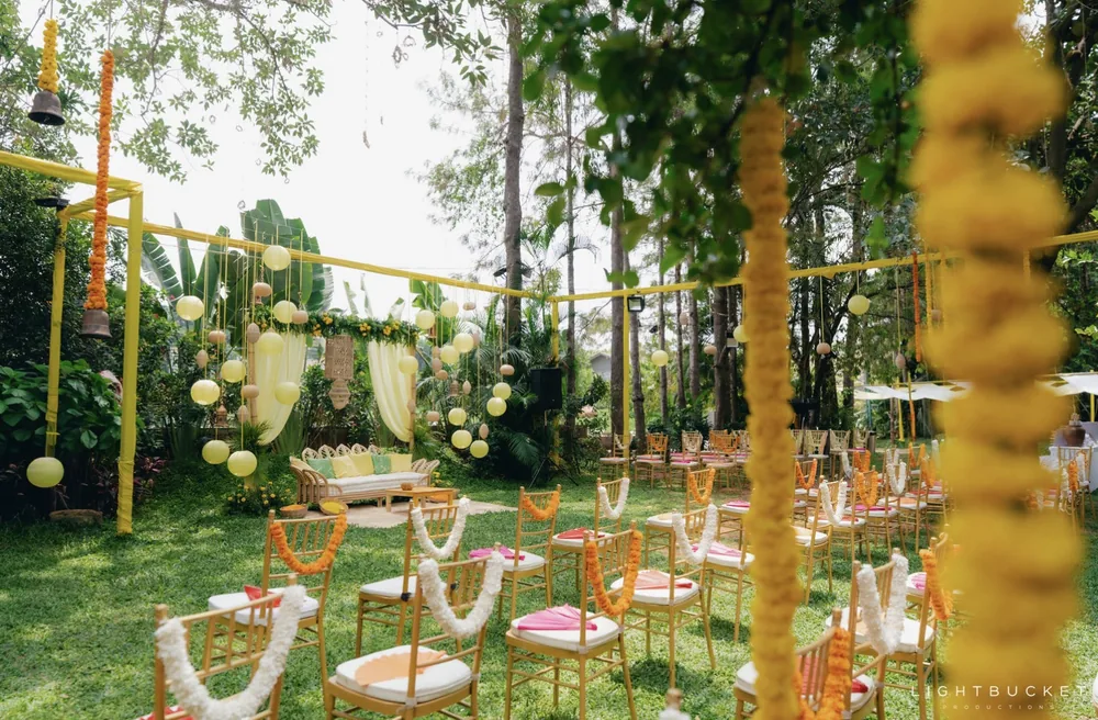 Garden lawn at Nature Knots, Bangalore, set for the haldi with gold Chiavari chairs, marigold garland strings and lanterns under tall trees