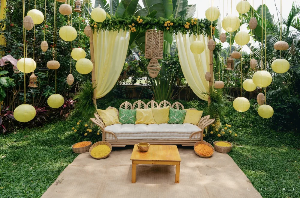 Wide view of the haldi lounge at Nature Knots, Bangalore: a cane sofa under sheer lemon drapes, paper lanterns, floor cushions and a low table on the lawn