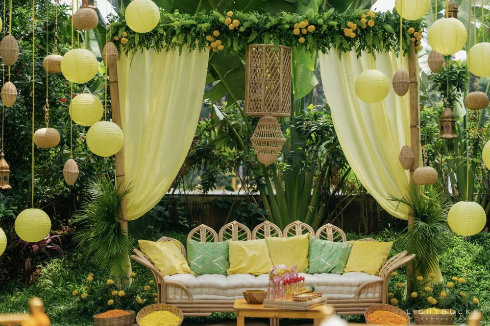 Lemon paper lanterns and wicker pendants hanging over the haldi sofa with green and yellow cushions at Nature Knots, Bangalore
