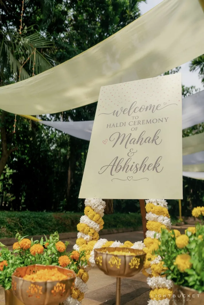 Haldi welcome board beside brass urns of marigold under sheer lemon canopies in the garden at Nature Knots, Bangalore