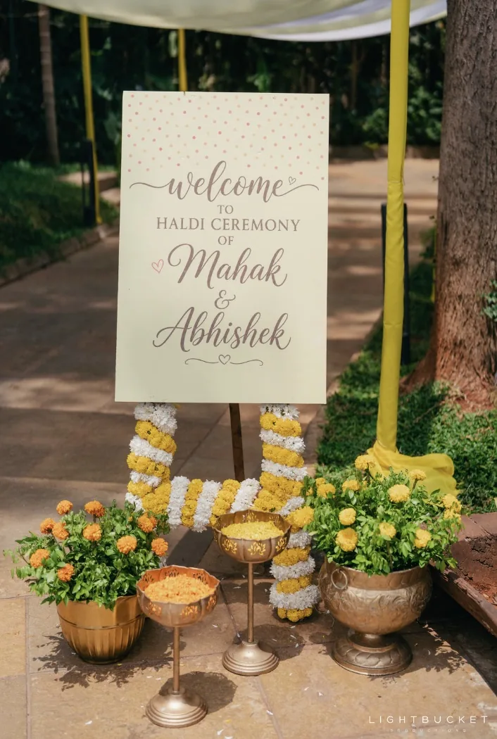 Haldi welcome board on an easel framed in jasmine and marigold garlands, with brass urns of marigold, at Nature Knots, Bangalore