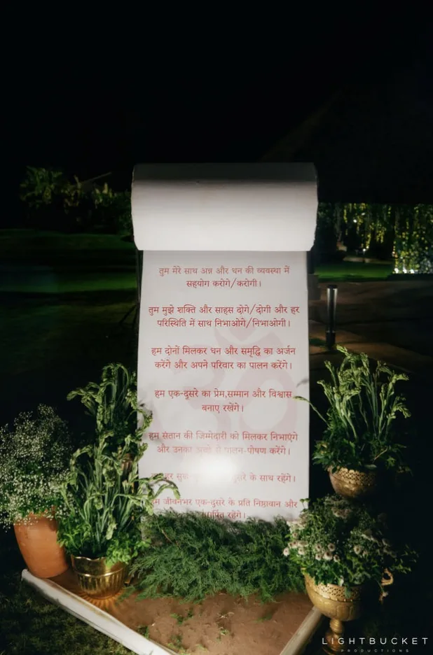 Freestanding scroll standee with the couple's wedding vows printed in Hindi among brass urns and plants on the lawn at Nature Knots, Bangalore