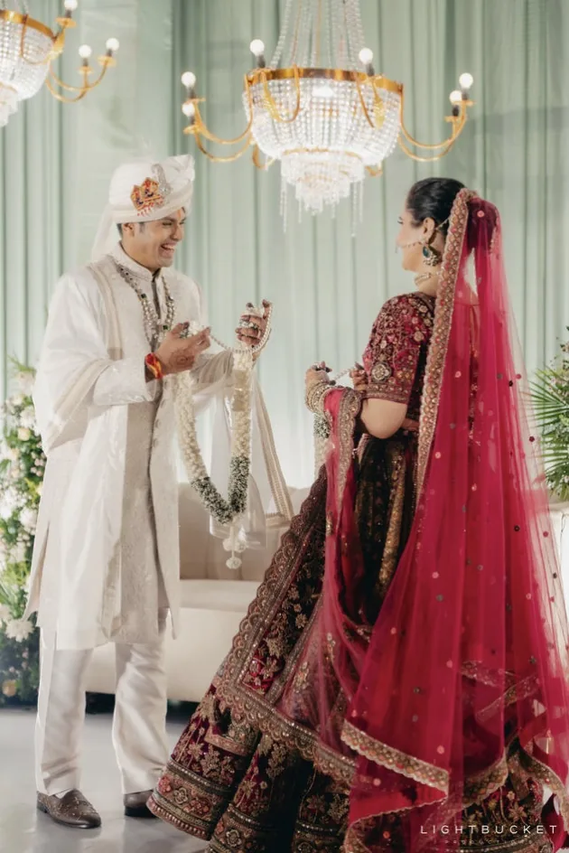 Couple exchanging jasmine jaimala garlands under crystal chandeliers against sage green drapes at Nature Knots, Bangalore