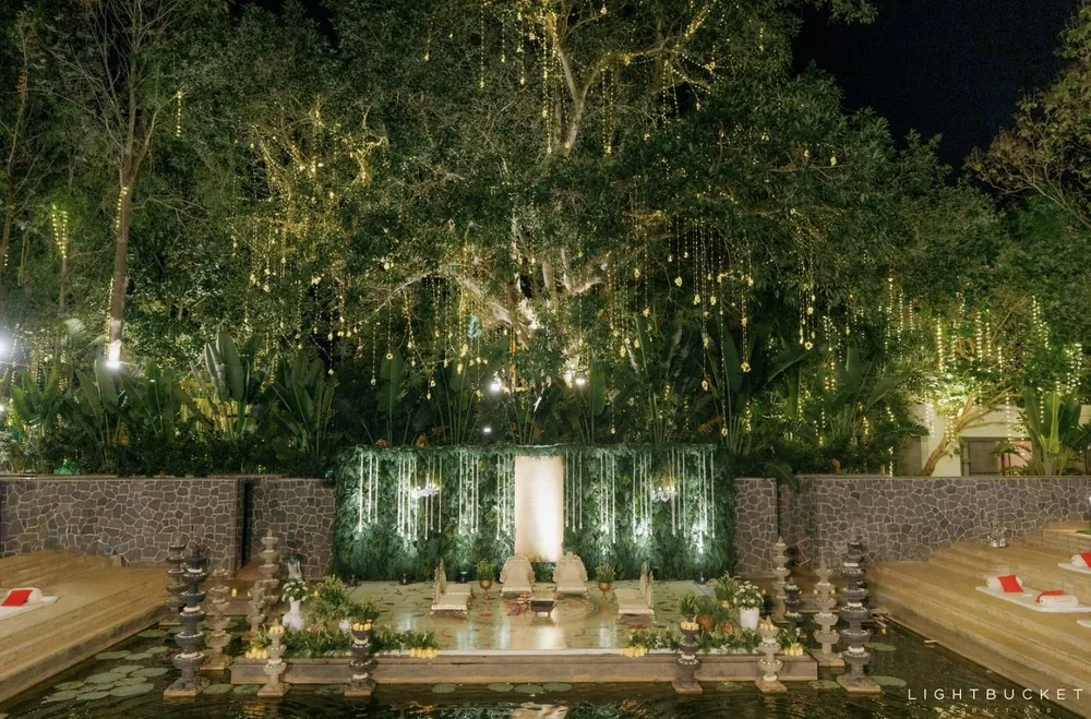 Wedding mandap on the lily pond at Nature Knots, Bangalore, under a banyan raining fairy lights, with stone lamp towers in the water and a glowing vows scroll