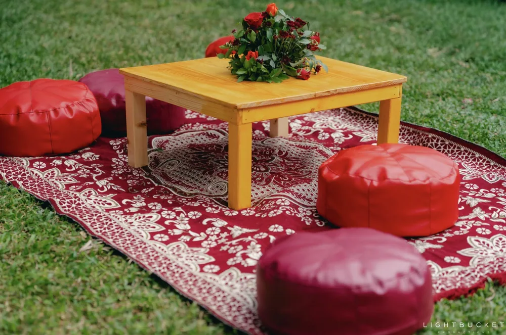 Red leather poufs around a low wooden table with a posy of red flowers on a patterned rug at the mehendi at Nature Knots, Bangalore