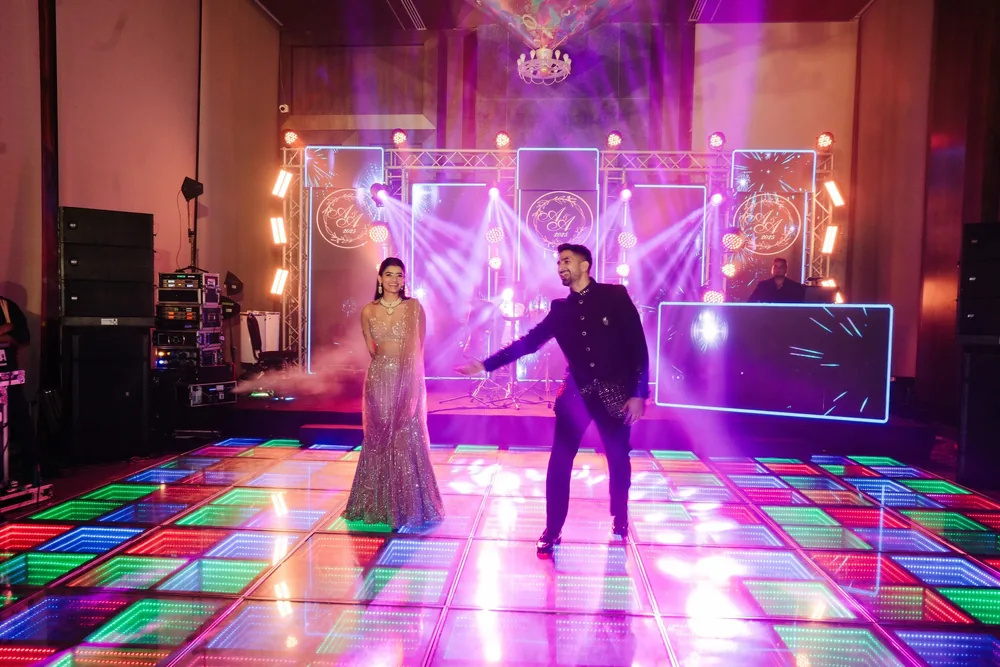 Apeksha and Akash dance on a multicolour LED floor at the after-party at Riva Beach Resort, Goa, before a DJ truss of monogram light panels in purple haze.