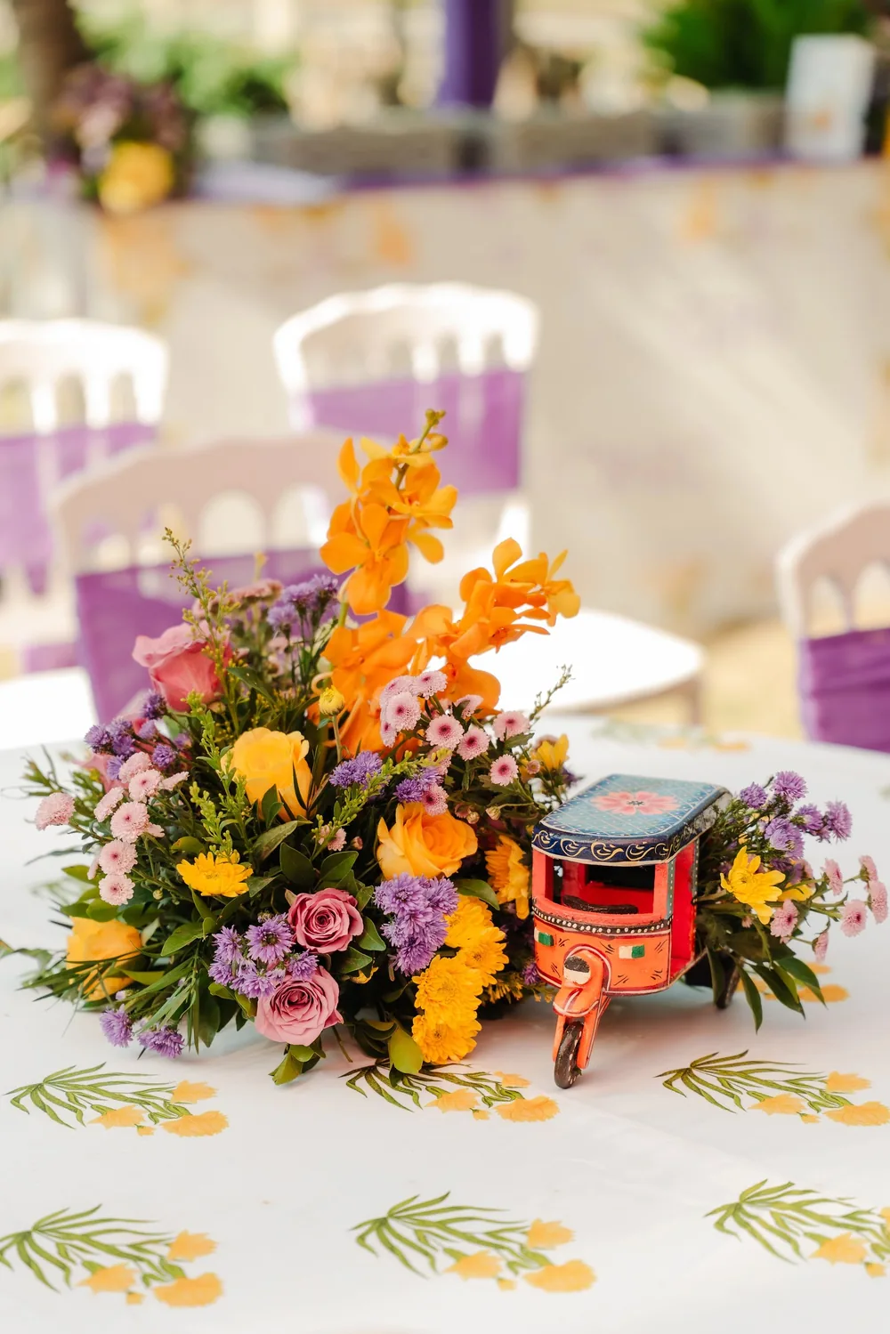 Haldi table centrepiece at Riva Beach Resort, Goa: orange orchids, roses and purple blooms beside a painted toy auto-rickshaw on a leaf-print tablecloth.
