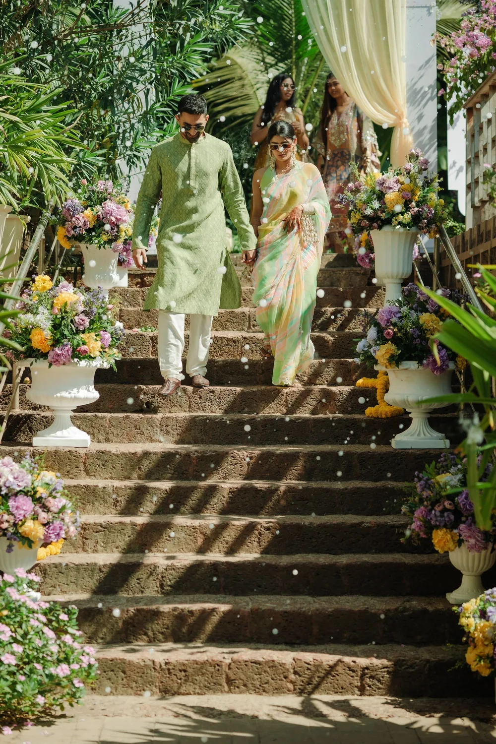 Apeksha and Akash walk down stone steps to the haldi at Riva Beach Resort, Goa, lined with white urns of lilac, yellow and pink flowers, a sheer drape above.