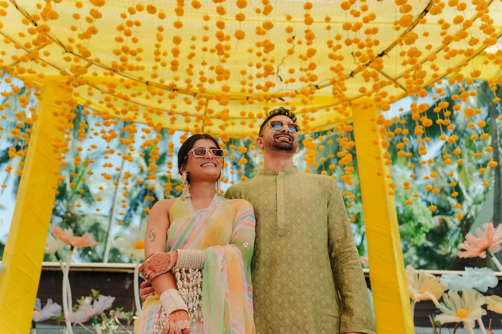 Apeksha and Akash in sunglasses laugh beneath the haldi canopy at Riva Beach Resort, Goa, with marigold strings overhead and giant pastel flowers behind.