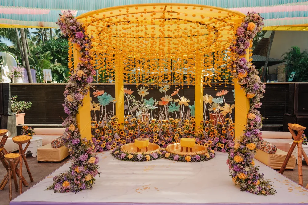 Haldi stage at Riva Beach Resort, Goa: a yellow canopy hung with marigold strings, lilac-and-yellow floral pillars, sunflowers and giant pastel blooms.