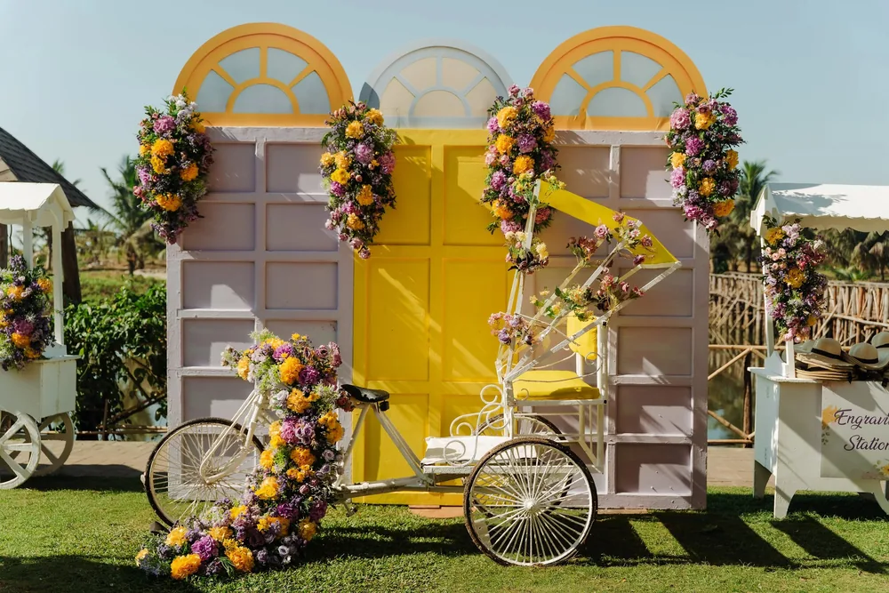 Haldi photobooth at Riva Beach Resort, Goa: lilac panels with a yellow door and arched windows, purple and orange flower clusters and a white cycle rickshaw.