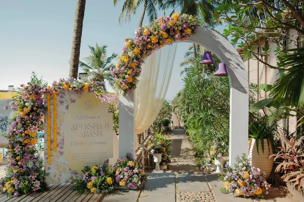 Haldi entrance at Riva Beach Resort, Goa: a white arch crowned with lilac, yellow and pink flowers, a sheer drape, purple hanging bells and a welcome sign.