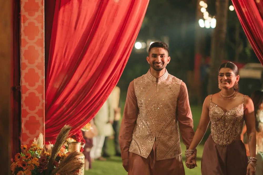 Apeksha and Akash walk hand in hand through red drapes into the mehendi at Riva Beach Resort, Goa, past orange flowers, pampas grass and garden lights.